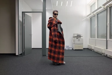 Model wearing an oversized plaid hooded coat inside a modern office space with dark carpet flooring.