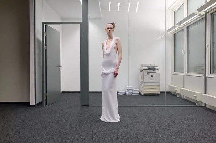 Model standing in a modern office wearing a long white sleeveless gown with feathered details