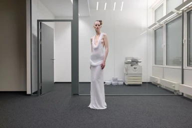 Model standing in a modern office wearing a long white sleeveless gown with feathered details