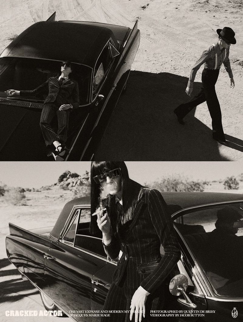 Black and white fashion shoot of a woman smoking next to a vintage car, with a man walking in the desert landscape.