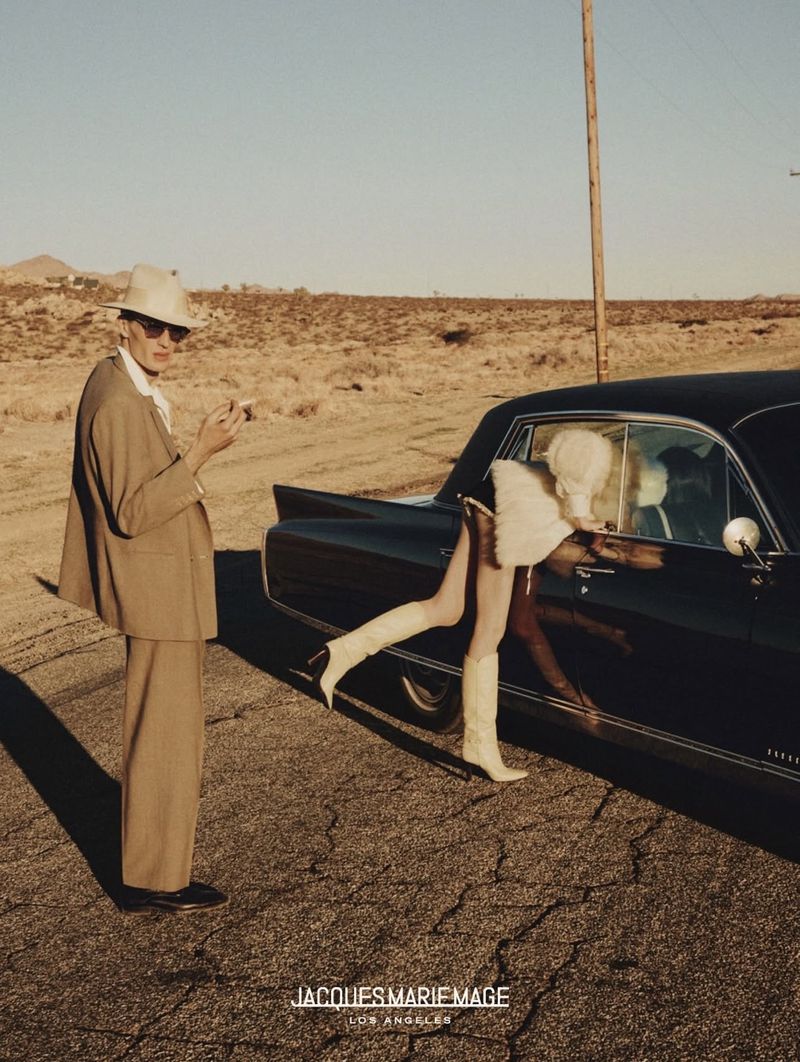 Man in a suit standing by a classic car with a woman in the window in a desert landscape under a clear sky during the day.
