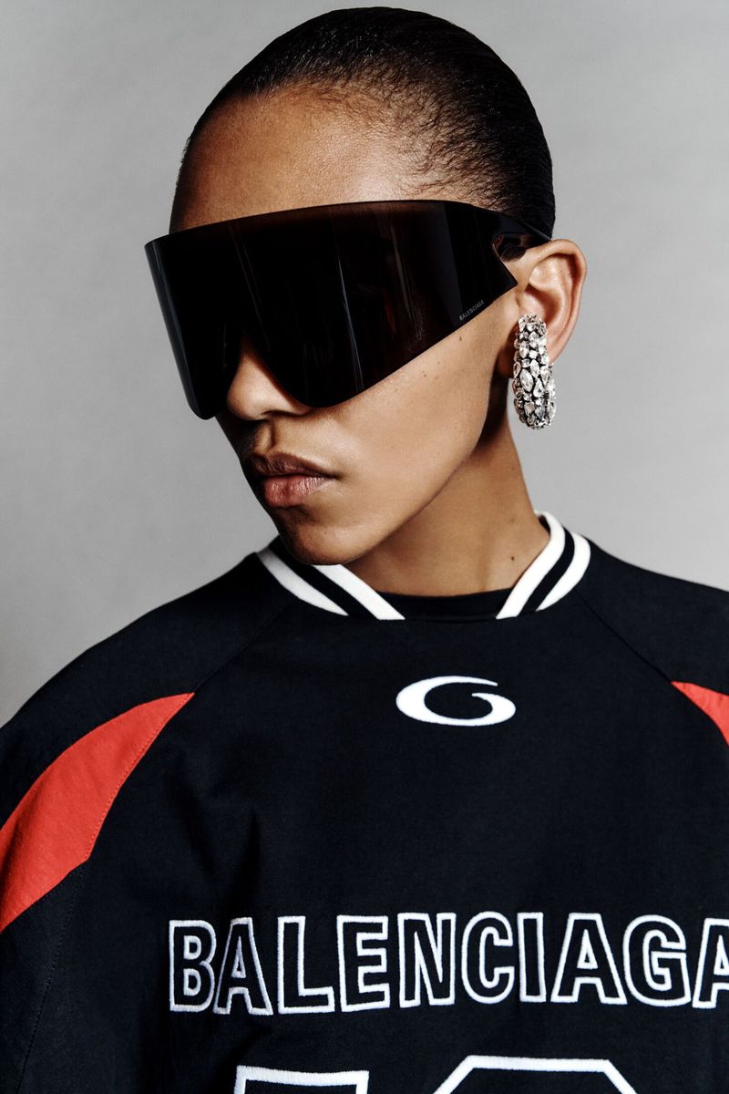 Woman with short hair wearing black sport jersey, oversized visor sunglasses, and rhinestone hoop earrings in a studio shot.
