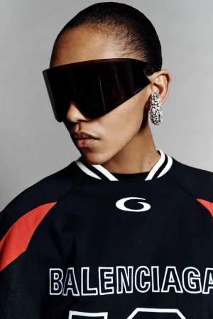 Woman with short hair wearing black sport jersey, oversized visor sunglasses, and rhinestone hoop earrings in a studio shot.