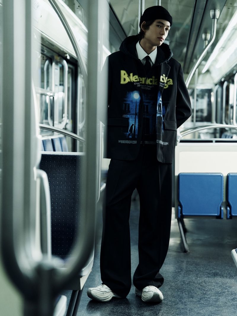 Young adult male stands in subway train aisle, wearing dark suit over hoodie with graphic, beanie, and white sneakers, fashion shot.
