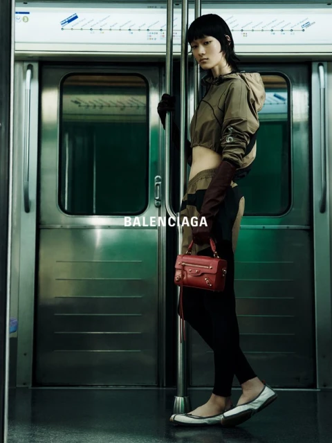Fashion shot of a woman in athletic wear holding a red purse while standing on a subway car, with metal doors and a subway map in view.