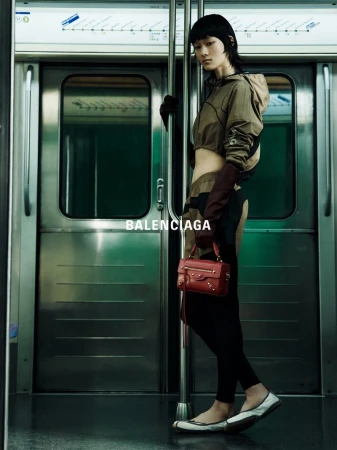 Fashion shot of a woman in athletic wear holding a red purse while standing on a subway car, with metal doors and a subway map in view.
