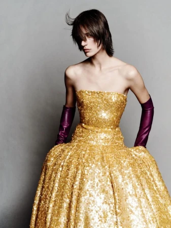 A woman in a strapless, gold, sequined gown stands in front of a grey background with dark hair and long, dark purple gloves.