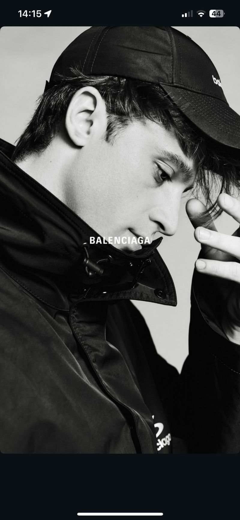 Monochrome portrait of a young adult man in a baseball cap and jacket, looking down with a hand near his face.