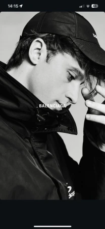 Monochrome portrait of a young adult man in a baseball cap and jacket, looking down with a hand near his face.