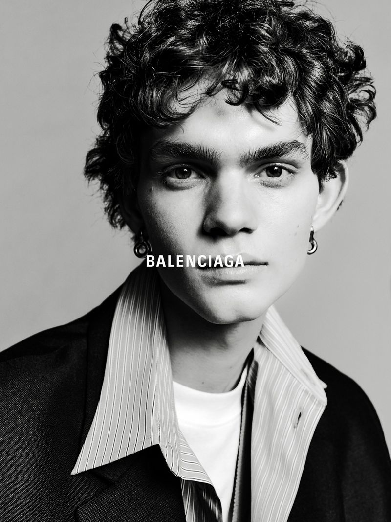 Black and white portrait of a young adult male wearing a blazer and striped shirt, featuring the Balenciaga logo near his neck.