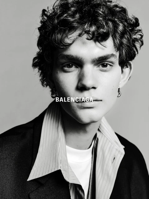 Black and white portrait of a young adult male wearing a blazer and striped shirt, featuring the Balenciaga logo near his neck.