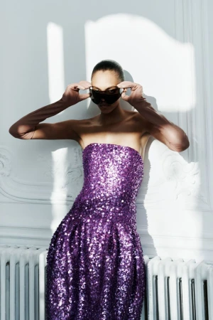 Woman in purple sequin dress and sunglasses poses in front of a white wall with soft shadows, creating a fashion editorial image.