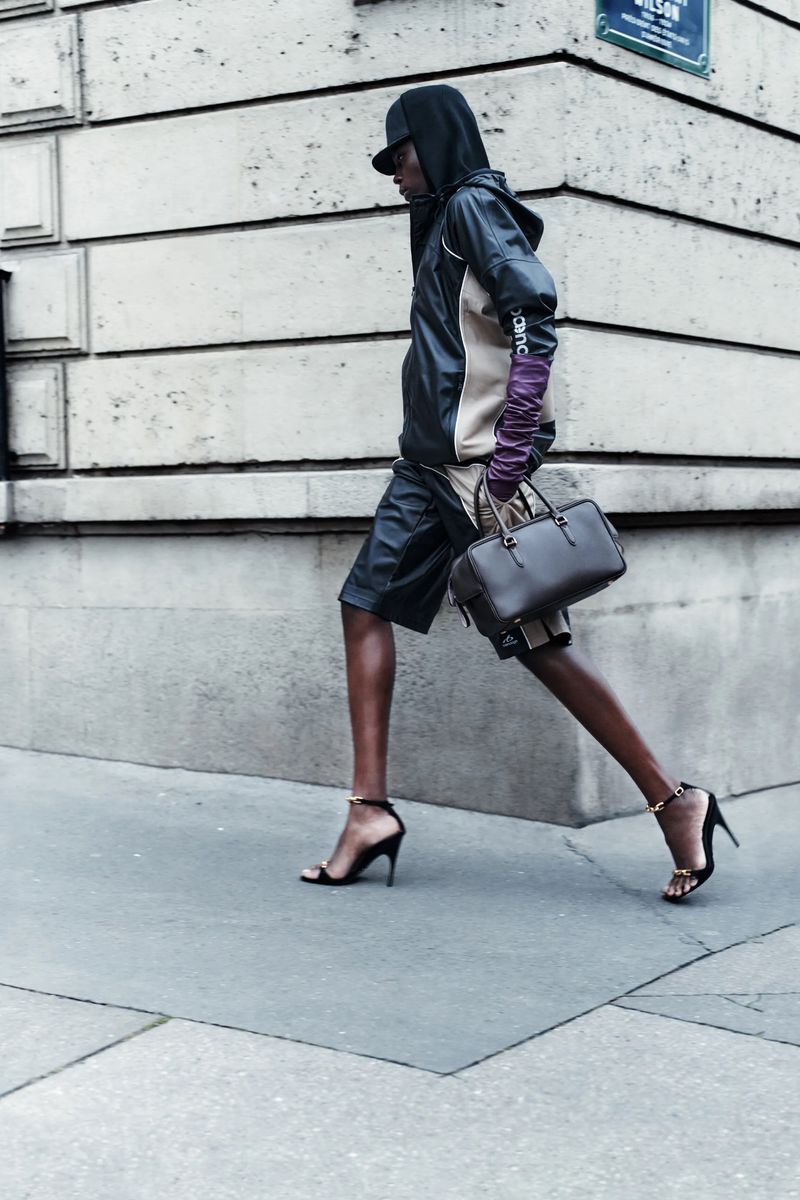 Fashionable adult model walks on city sidewalk wearing hoodie, leather shorts, high heels, long gloves, and carrying a dark handbag.