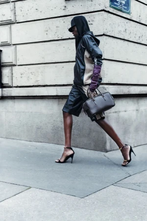 Fashionable adult model walks on city sidewalk wearing hoodie, leather shorts, high heels, long gloves, and carrying a dark handbag.