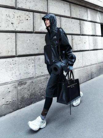 A person walks along a city street wearing a black jacket, jeans, and sneakers, carrying a black tote bag and yoga mat.