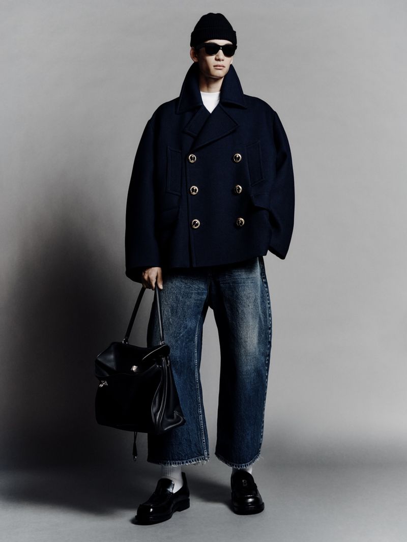 Man in navy peacoat, black beanie, and sunglasses carrying a black leather bag. Wide-leg jeans and loafers. Full studio shot.