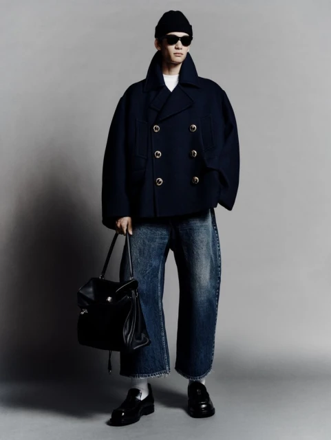 Man in navy peacoat, black beanie, and sunglasses carrying a black leather bag. Wide-leg jeans and loafers. Full studio shot.