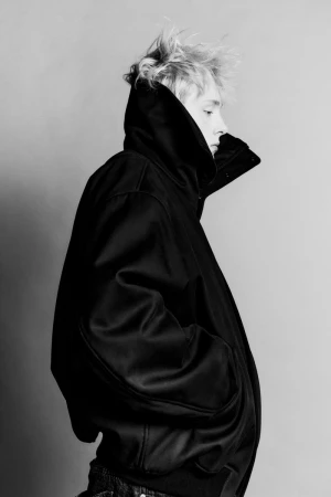 Monochrome portrait of a person with blonde hair wearing a black hooded jacket, captured in a studio setting with soft lighting.