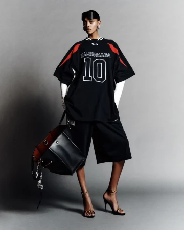 Studio shot of a Black fashion model wearing a Balenciaga shirt, high heels, and carrying a leather bag, showcasing contemporary style.
