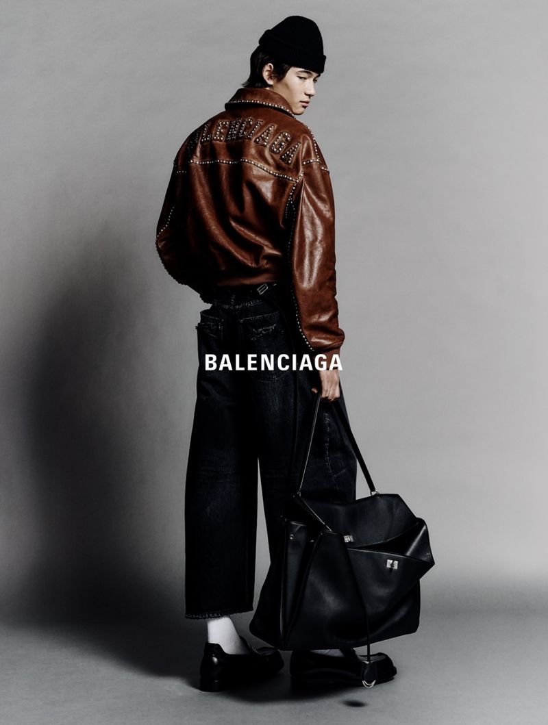 Young adult male poses in a studio wearing a brown leather jacket, black jeans, a black beanie, and carrying a large black tote bag.