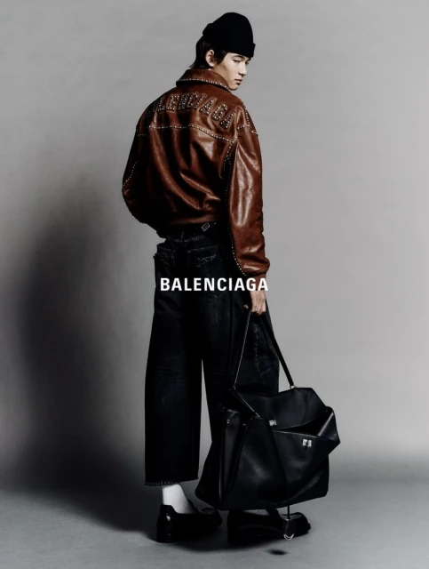 Young adult male poses in a studio wearing a brown leather jacket, black jeans, a black beanie, and carrying a large black tote bag.