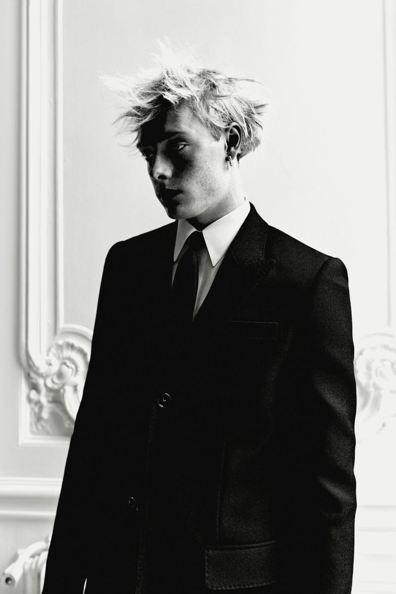 Black and white portrait of a blonde man in a dark suit, white shirt, and black tie against a decorative wall. Fashion editorial.