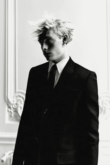 Black and white portrait of a blonde man in a dark suit, white shirt, and black tie against a decorative wall. Fashion editorial.