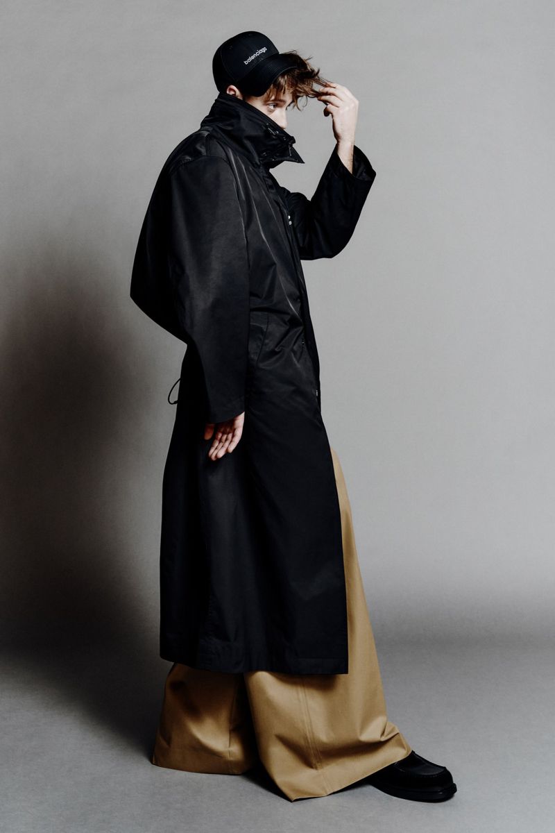 Person in a black rain coat, black baseball cap, and wide tan pants standing in a photo studio against a neutral gray background.