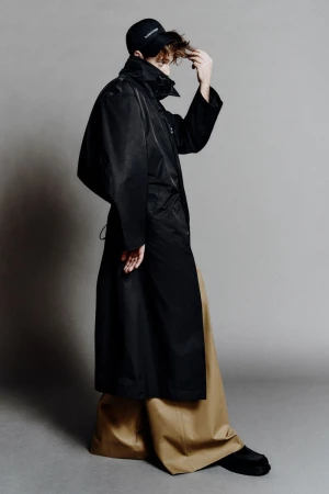 Person in a black rain coat, black baseball cap, and wide tan pants standing in a photo studio against a neutral gray background.