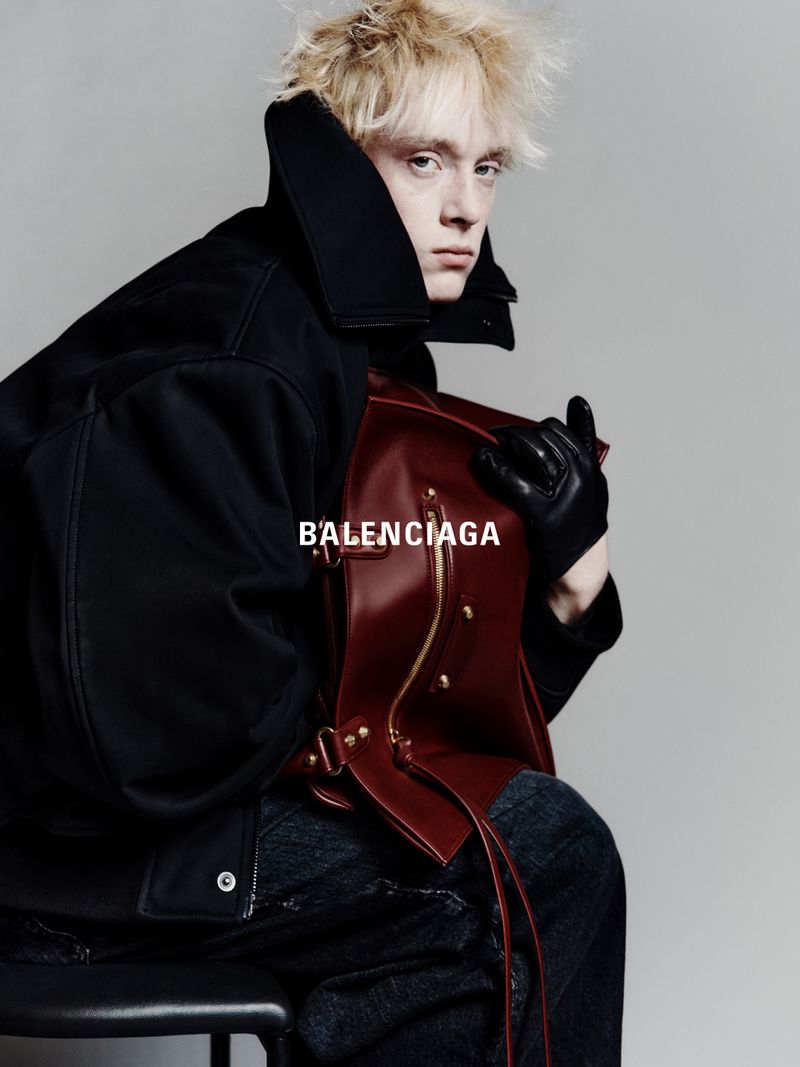 Fashion shot of a blonde adult male in a dark coat holding a leather bag while sitting on a stool against a grey backdrop, Balenciaga logo.