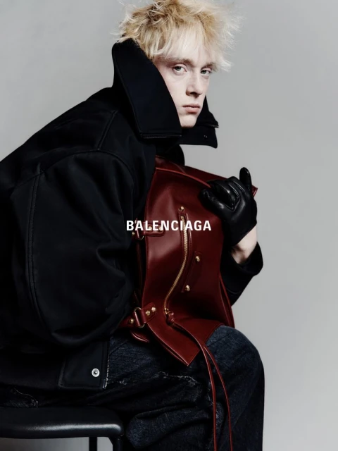 Fashion shot of a blonde adult male in a dark coat holding a leather bag while sitting on a stool against a grey backdrop, Balenciaga logo.
