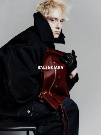 Fashion shot of a blonde adult male in a dark coat holding a leather bag while sitting on a stool against a grey backdrop, Balenciaga logo.