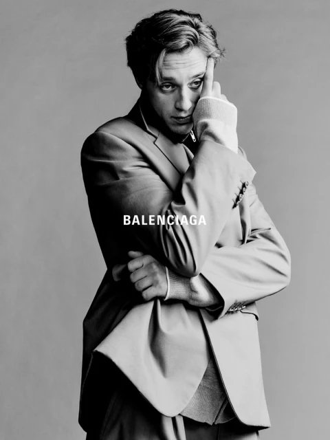 Monochrome studio shot of a young adult man in a suit, posing with his hand to his forehead; Balenciaga logo is visible on the suit.