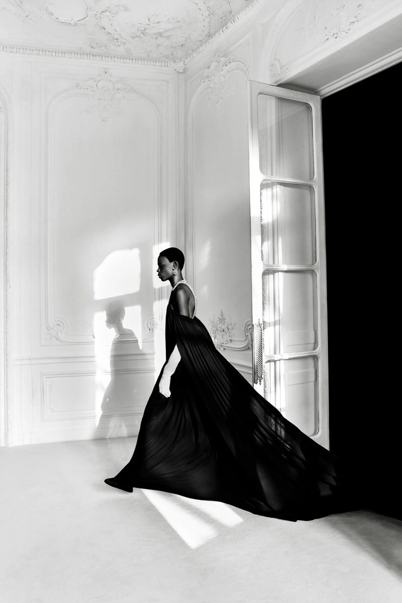 Elegant fashion shot: a Black woman in a flowing black gown walks in a white room with a large doorway and soft natural light.