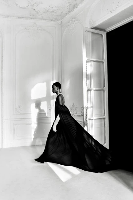 Elegant fashion shot: a Black woman in a flowing black gown walks in a white room with a large doorway and soft natural light.