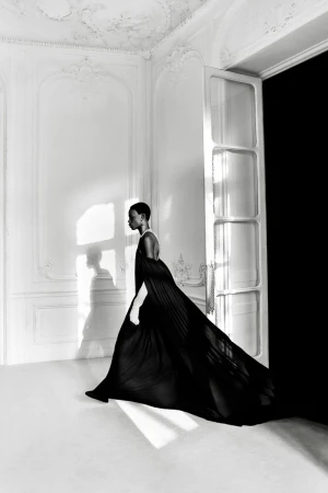 Elegant fashion shot: a Black woman in a flowing black gown walks in a white room with a large doorway and soft natural light.