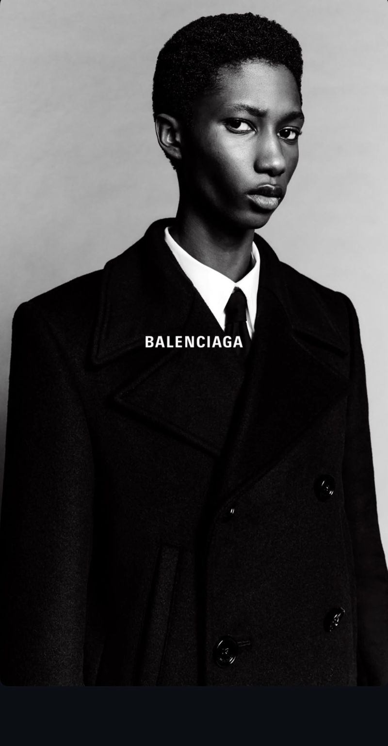 Elegant androgynous model with dark hair and skin, in a black coat, shirt and tie, poses in a fashion shot with 'BALENCIAGA' text.