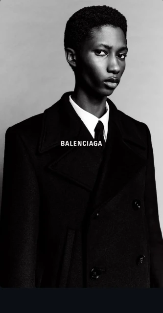 Elegant androgynous model with dark hair and skin, in a black coat, shirt and tie, poses in a fashion shot with 'BALENCIAGA' text.