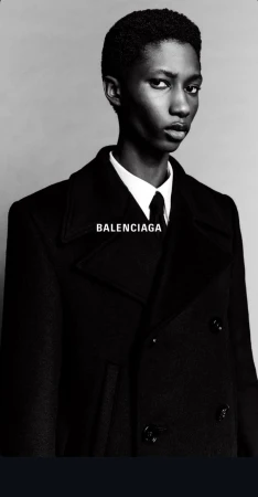 Elegant androgynous model with dark hair and skin, in a black coat, shirt and tie, poses in a fashion shot with 'BALENCIAGA' text.
