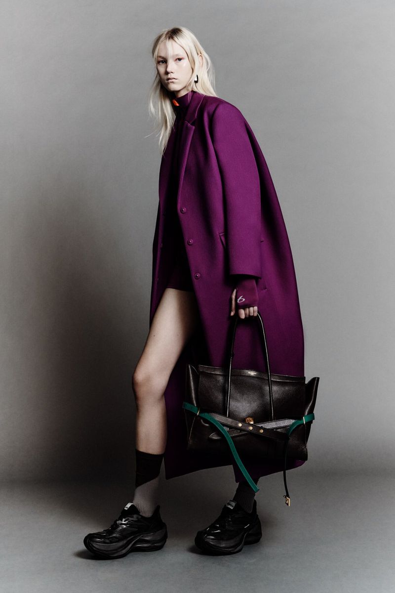 Woman in a magenta coat holds a black tote bag, showcasing high-fashion against a plain studio backdrop with edgy socks and platform shoes.