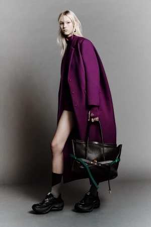 Woman in a magenta coat holds a black tote bag, showcasing high-fashion against a plain studio backdrop with edgy socks and platform shoes.