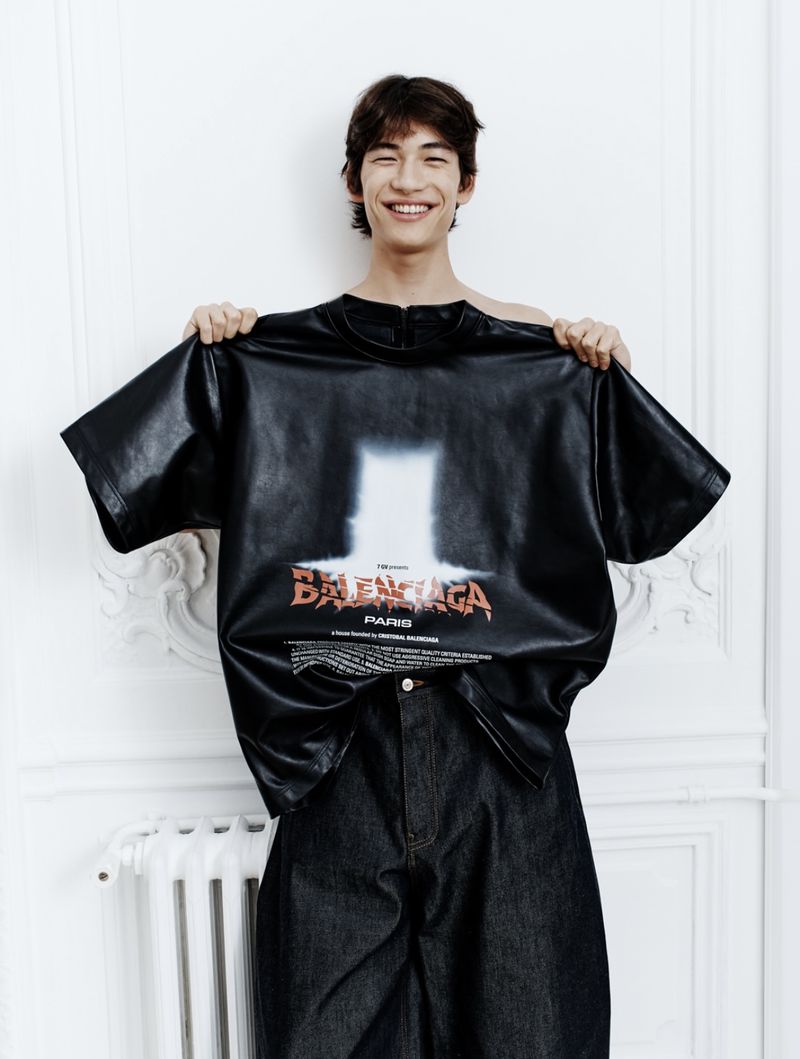 Young man in an indoor setting holds up a Balenciaga t-shirt with graphic design, posing for a fashion editorial-style shot.