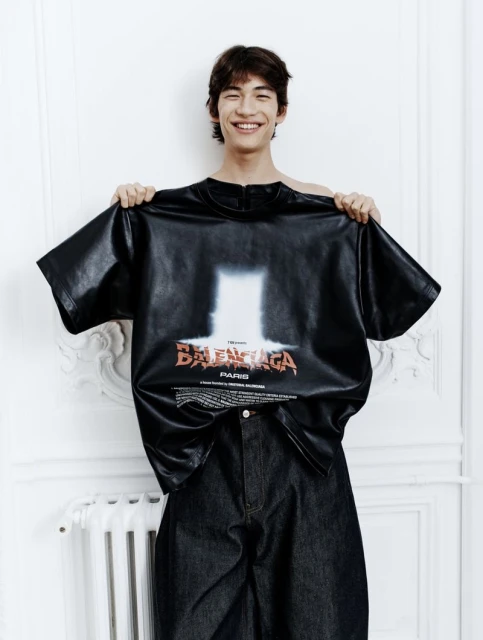 Young man in an indoor setting holds up a Balenciaga t-shirt with graphic design, posing for a fashion editorial-style shot.