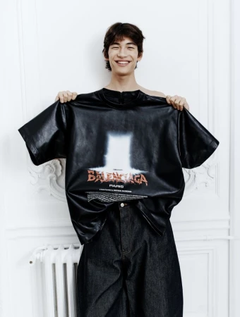 Young man in an indoor setting holds up a Balenciaga t-shirt with graphic design, posing for a fashion editorial-style shot.