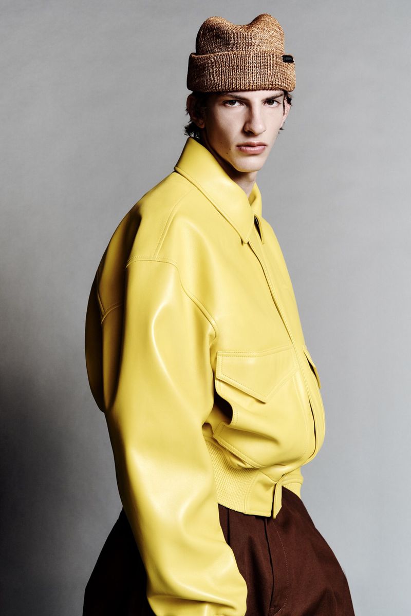 Portrait of a male fashion model in a yellow leather jacket and knit hat posing in a studio with a neutral grey backdrop.