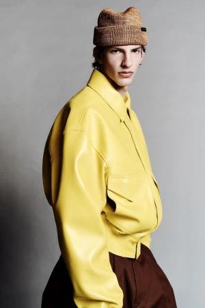 Portrait of a male fashion model in a yellow leather jacket and knit hat posing in a studio with a neutral grey backdrop.