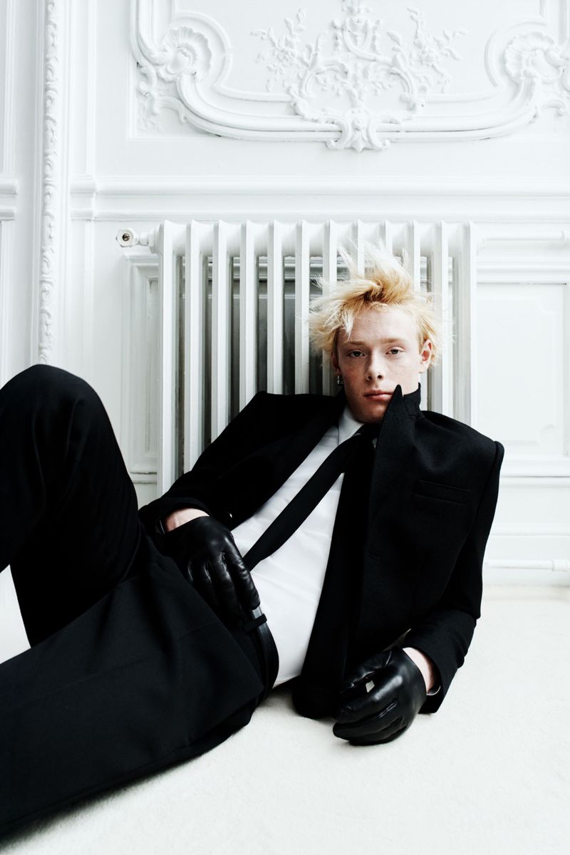 A stylish adult male with light blond hair is reclining in a fashionable black suit with gloves, indoors against a white ornate wall.