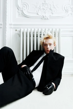 A stylish adult male with light blond hair is reclining in a fashionable black suit with gloves, indoors against a white ornate wall.