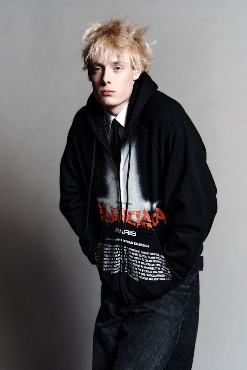 Portrait of a young adult male with blond hair wearing a black printed zip-up hoodie, white collared shirt, and dark jeans.