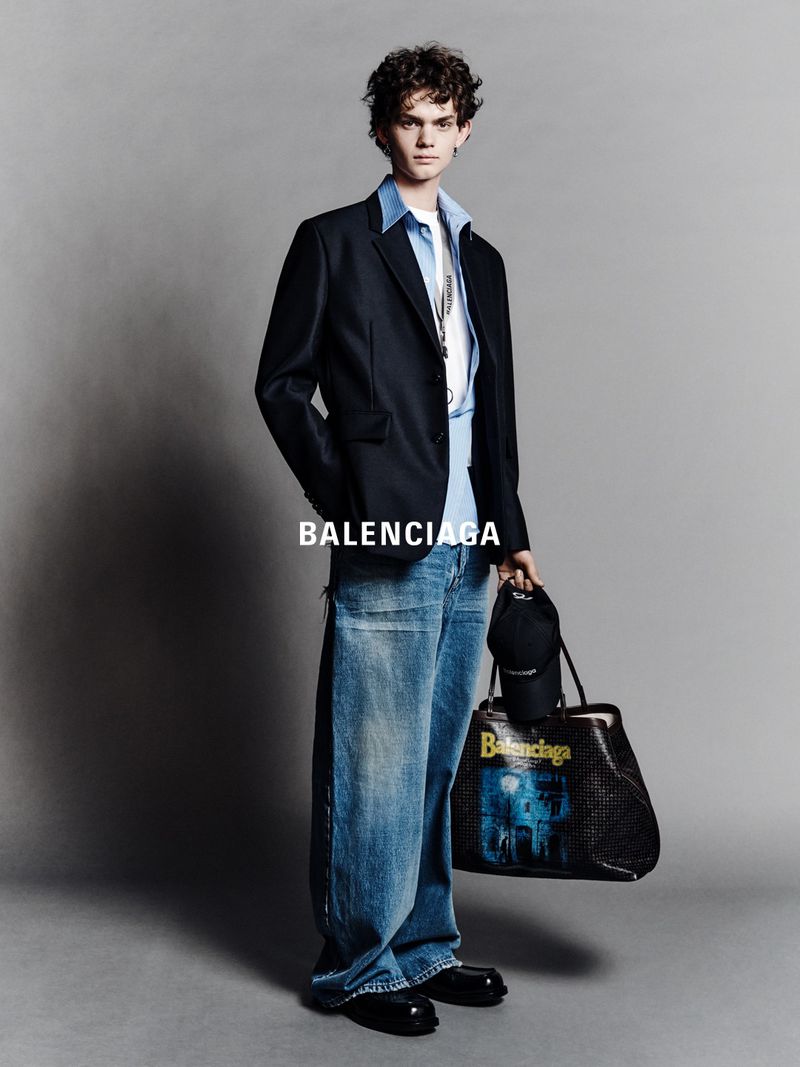 Full-length portrait of a young adult male wearing a black blazer, light blue button-down shirt, baggy jeans, and carrying a tote bag.